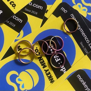 Puede incluir: Una colección de pequeños anillos metálicos iridiscentes en tonos rosa y dorado, dispersos sobre un fondo de tarjetas amarillas y azules. Las tarjetas tienen el texto "monkeymenco.co" y "Since 2018".