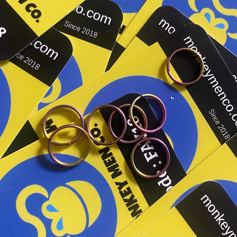 May include: A collection of small, metallic rings in shades of pink and gold, scattered on a background of yellow and blue cards. The cards feature the text "monkeymenco.co" and "Since 2018".
