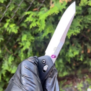 Könnte beinhalten: Ein silbernes Taschenmesser mit schwarzem Griff und einem violetten Akzent an der Pivot-Schraube. Das Messer ist teilweise geöffnet und die Klinge ist sichtbar.
