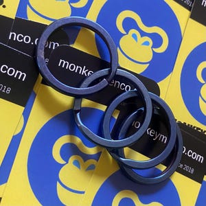 May include: Four linked, metallic blue key rings. The rings are circular and have a smooth finish. The background features business cards with a yellow and blue monkey logo and the text "monkeyenco.com".