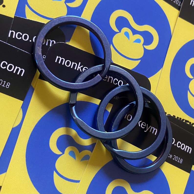 May include: Four linked, metallic blue key rings. The rings are circular and have a smooth finish. The background features business cards with a yellow and blue monkey logo and the text "monkeyenco.com".