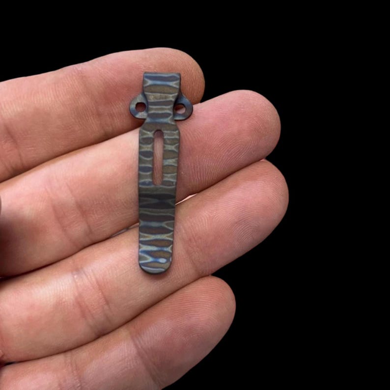 May include: A dark grey and blue patterned metal pocket clip with two holes at the top and a slot in the middle.