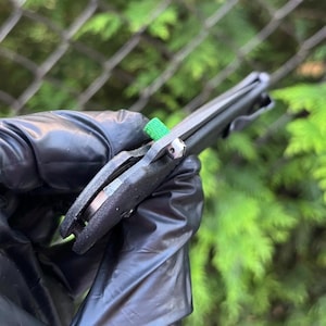 Könnte beinhalten: Ein schwarzes Klappmesser mit einem grünen Akzent am Griff. Das Messer wird in einer schwarzen, behandschuhten Hand gehalten.
