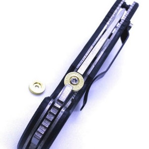May include: A black folding knife with a silver blade and a gold-colored bullet casing on the handle. The knife is closed and the bullet casing is visible.