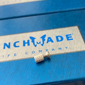 Könnte beinhalten: Eine silberne Metallschraube mit einem gerändelten Kopf, die auf einem blauen Kasten mit dem aufgedruckten Text "Benchmade Company" liegt.