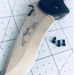 May include: A silver Emerson Wave knife with a black handle and a bottle opener. The knife has the text "M-COM MADE IN USA" and "EMERSON WAVE FEATURE US PAT NO 5878500" on the blade. The knife is on a white grid background with three black screws.