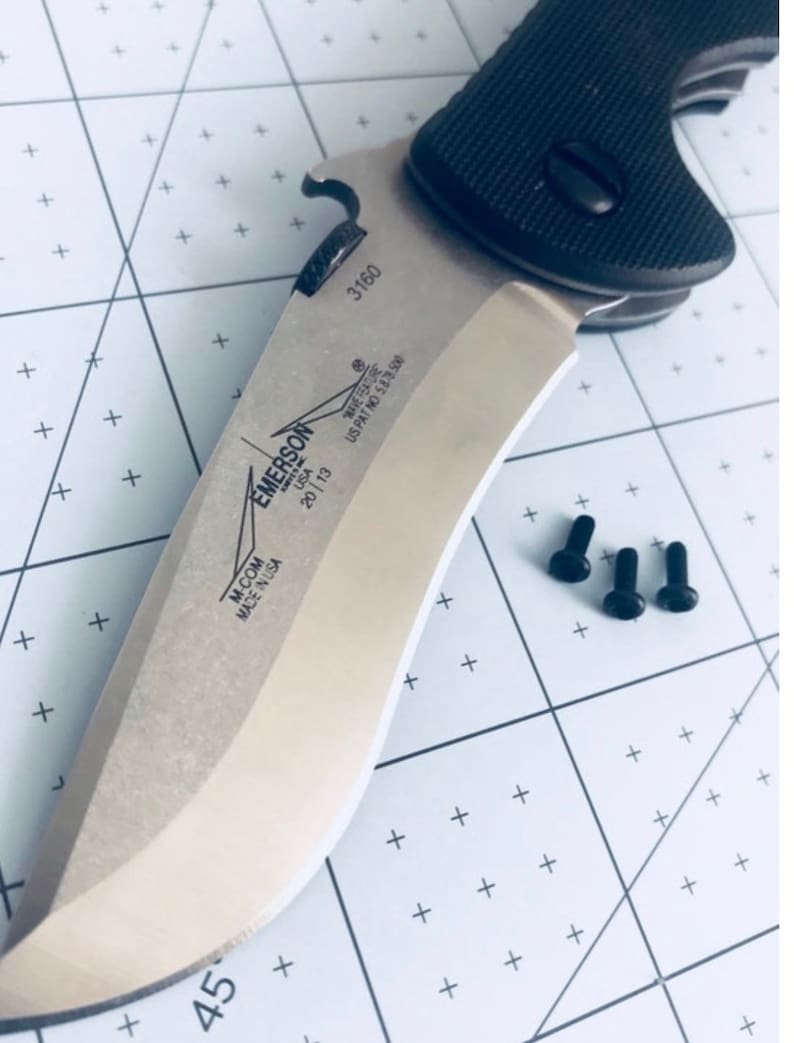 May include: A silver Emerson Wave knife with a black handle and a bottle opener. The knife has the text "M-COM MADE IN USA" and "EMERSON WAVE FEATURE US PAT NO 5878500" on the blade. The knife is on a white grid background with three black screws.