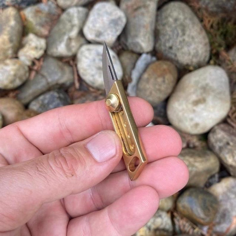 May include: A small, gold-colored pocket knife with a sharp, silver blade. The knife is held in a hand, with the blade extended. The knife is made of metal and has a simple, elegant design. The background is out of focus, with rocks and pebbles.