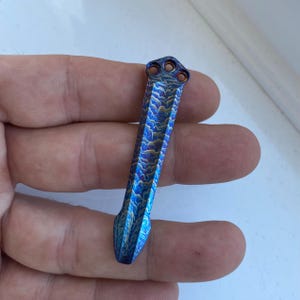 May include: A blue and gold titanium pocket tool with a unique wave pattern. The tool has a pointed tip and a rounded top with three holes.