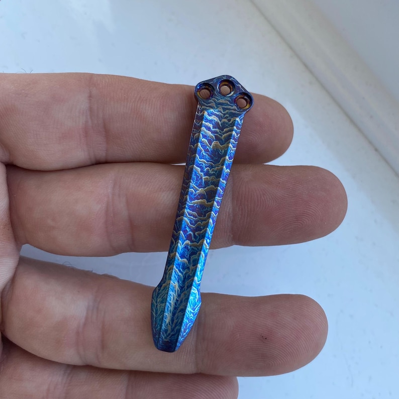 May include: A blue and gold titanium pocket tool with a unique wave pattern. The tool has a pointed tip and a rounded top with three holes.