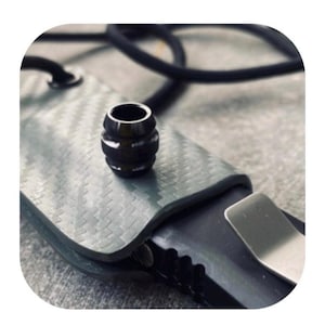 Puede incluir: Una cuenta negra cilíndrica con una superficie texturizada se encuentra sobre una funda gris texturizada. También se ven un cordón negro y un clip plateado. La cuenta es probablemente un elemento decorativo para un cuchillo o herramienta similar.