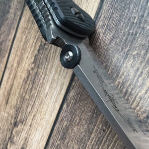 May include: A black and silver folding knife with a textured black handle and a partially open blade. The blade is labeled "Emerson CQC-7K" and "Made in USA".