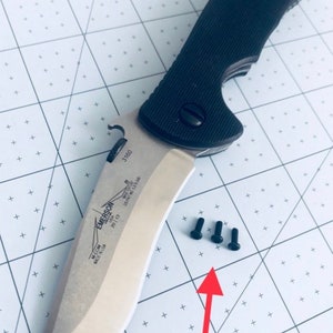 May include: A black and silver Emerson knife with a partially open blade. The knife has a black handle with a textured surface. Three small black screws are visible on a white surface.