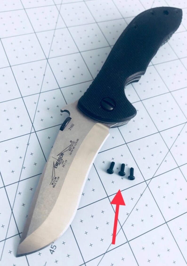 May include: A black and silver Emerson knife with a partially open blade. The knife has a black handle with a textured surface. Three small black screws are visible on a white surface.