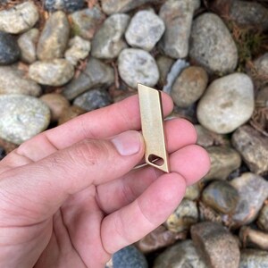 May include: A small, rectangular, gold-colored metal pendant with a cut-out oval shape near the bottom. The pendant is held against a background of rocks and foliage.