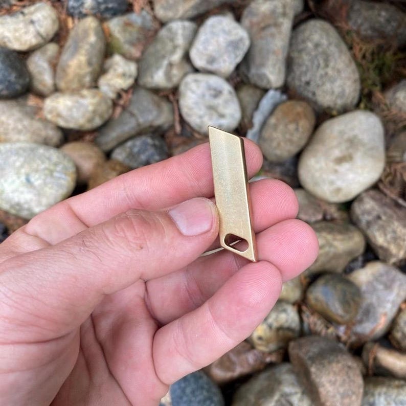 May include: A small, rectangular, gold-colored metal pendant with a cut-out oval shape near the bottom. The pendant is held against a background of rocks and foliage.
