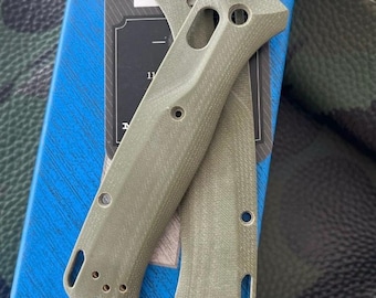May include: Two olive-green knife handle scales with multiple holes and a textured surface. The scales are designed to fit a folding knife. The background includes a blue box and a camouflage pattern.