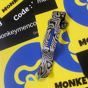 Peut inclure: Un clip de poche en métal à motifs avec un accent bleu. Le clip présente un motif ondulé et marbré dans des tons argent et noir. Le clip est posé sur un fond de cartes jaunes et bleues avec le mot "MONKEY" et le texte "Code: 1" visibles.