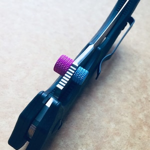 Könnte beinhalten: Ein schwarzes Klappmesser mit einer silbernen Klinge und einer pinkfarbenen und blauen Schraube am Griff.