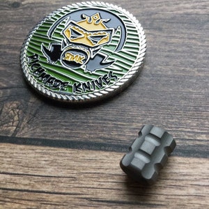 Puede incluir: Una moneda de plata con un gráfico verde y negro de una figura enmascarada con el texto "Richard Made Knives" alrededor del borde. También hay un pequeño fidget spinner de metal gris en la imagen.