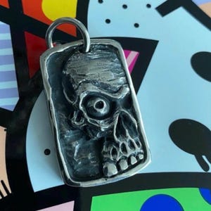 Op de afbeelding: Zilverkleurige rechthoekige hanger met een gedetailleerd schedelontwerp. De schedel heeft een prominente oogkas en zichtbare tanden. De hanger heeft een getextureerd oppervlak en een lus voor een ketting.
