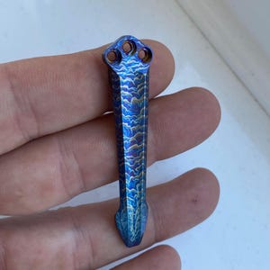 May include: A blue and gold titanium pocket knife with a wave pattern. The knife has a pointed tip and three holes at the top.