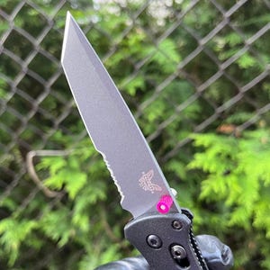 Könnte beinhalten: Ein schwarzes Klappmesser mit einer teilweise gezahnten Klinge und einem rosa Akzent am Griff. Das Messer hat ein Schmetterlingslogo auf der Klinge.