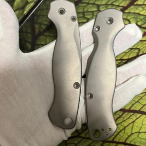 May include: Two silver knife handles with a curved design and multiple holes. The handles are displayed against a green and white patterned background, held by a gloved hand.