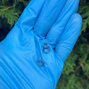 Puede incluir: Tres pequeños anillos metálicos de color plateado se muestran sobre un fondo azul. Dos anillos están conectados y uno está separado. Los anillos son circulares y parecen estar hechos de metal brillante.