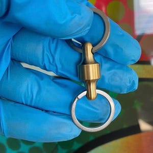 Könnte beinhalten: Ein Schlüsselanhänger aus Messing und Silber mit Magnetverschluss. Der Schlüsselanhänger hat zwei silberne Schlüsselringe, die durch eine zylindrische Messingkomponente verbunden sind. Das Messingteil hat einen Magnetverschluss. Der Hintergrund ist verschwommen, mit blauen und bunten Elementen.