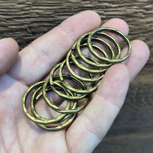 Peut inclure: Un ensemble de porte-clés de couleur laiton antique. Les anneaux ont une texture martelée et sont empilés dans une main. Les anneaux sont de couleur dorée chaude.