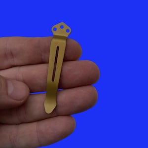 Pu&ograve; includere: Un fermaglio da tasca in metallo color oro con una parte superiore arrotondata e un bordo dritto. Il fermaglio ha tre piccoli fori nella parte superiore.