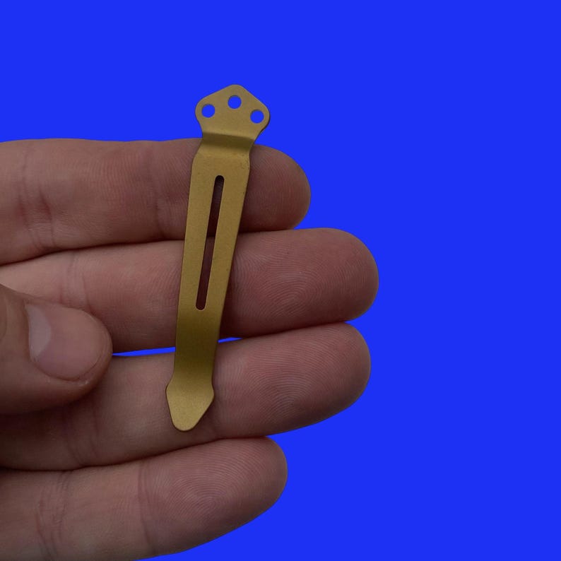 Pu&ograve; includere: Un fermaglio da tasca in metallo color oro con una parte superiore arrotondata e un bordo dritto. Il fermaglio ha tre piccoli fori nella parte superiore.