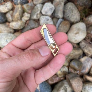 May include: A small, gold-colored metal tool with a geometric design. It features a silver and gold blade and a circular element. The tool is held in a hand against a background of rocks.