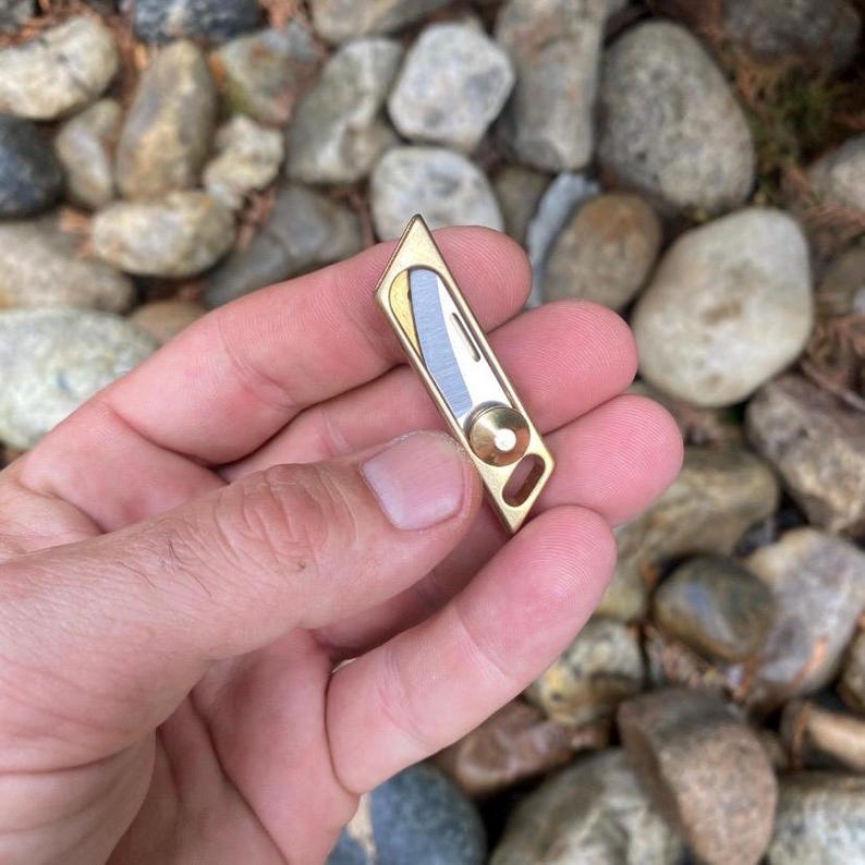 May include: A small, gold-colored metal tool with a geometric design. It features a silver and gold blade and a circular element. The tool is held in a hand against a background of rocks.