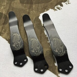 May include: Three black metal pocket clips with a decorative engraved design of a koi fish. Each clip has four holes for attaching to a pocket.