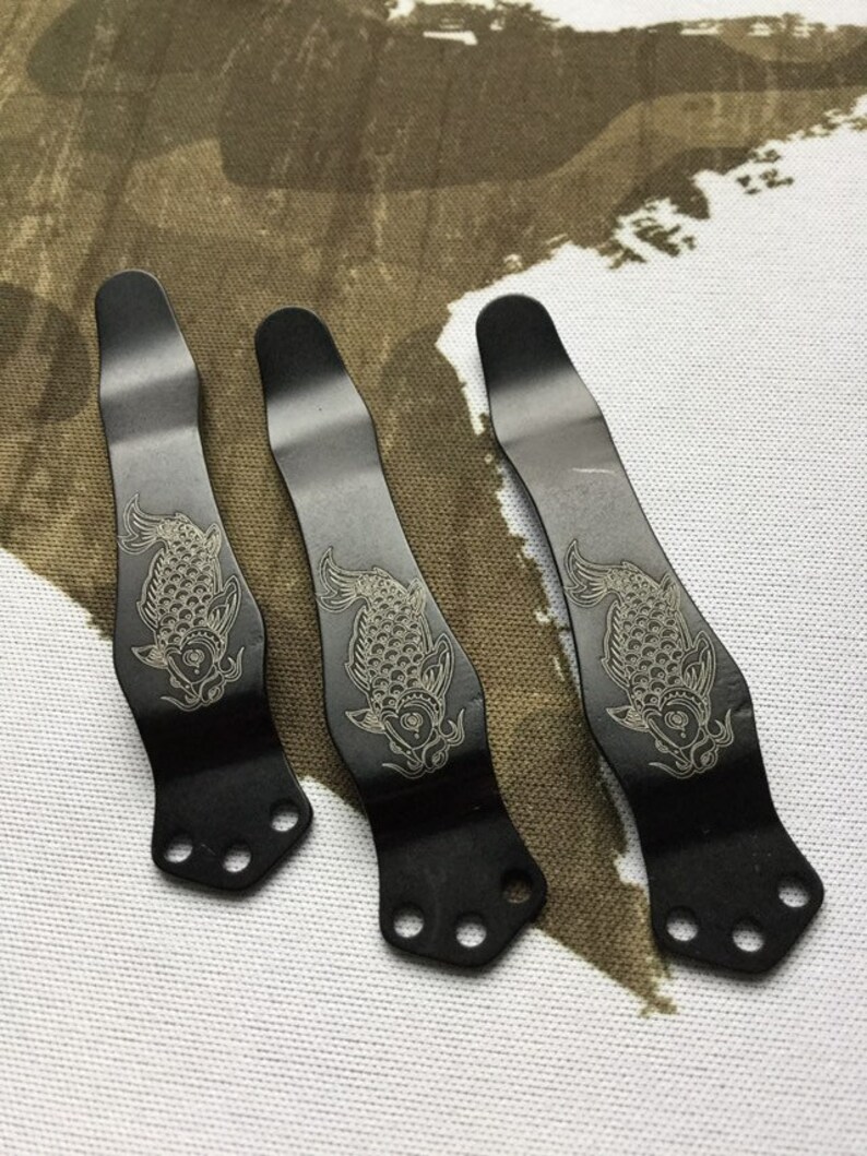 May include: Three black metal pocket clips with a decorative engraved design of a koi fish. Each clip has four holes for attaching to a pocket.