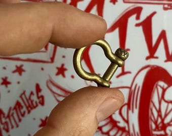 May include: A small, brass-colored shackle held between two fingers. The shackle has a D-shape with a screw pin closure. The background is a white surface with red star and text designs.