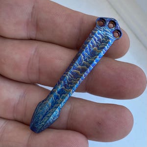 May include: A blue and gold titanium pocket tool with a wavy pattern. The tool has a rounded tip and a hole at the top.