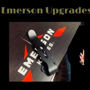 May include: A black pocket clip for a knife, with a silver top, is shown against a dark background. The text "Emerson Knives" is visible in white, with red diamond accents. The words "Emerson Upgrades" are at the top.