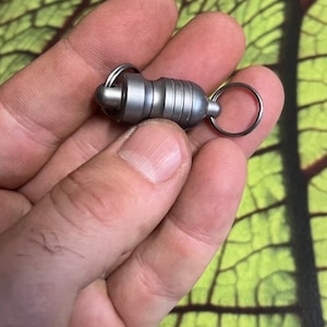Könnte beinhalten: Ein silberner Schlüsselanhänger aus Metall mit einem magnetischen Verschluss und einem kleinen Ring. Der Schlüsselanhänger wird in einer Hand gehalten.
