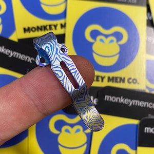 Peut inclure: Un clip de poche en métal multicolore avec un motif ondulé unique. Le clip est présenté sur un fond d'autocollants jaunes et bleus avec un logo de singe et le texte "MONKEY MEN co."