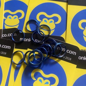 May include: A collection of small, blue metal rings scattered over yellow cards with a blue monkey logo. The cards also feature the text "keymenco.com Since 2018". The rings appear to be for jewelry or crafts.