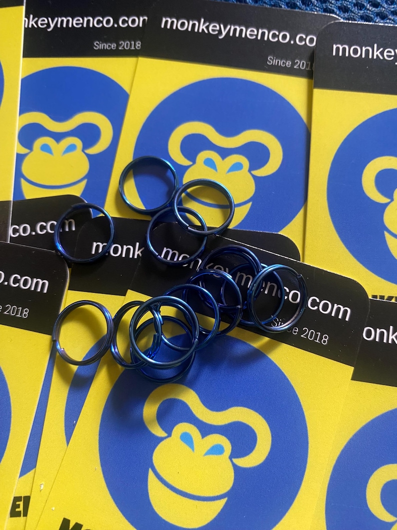 May include: A collection of small, blue metal rings scattered over yellow cards with a blue monkey logo. The cards also feature the text "keymenco.com Since 2018". The rings appear to be for jewelry or crafts.
