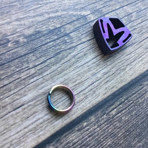 May include: A small, iridescent ring and a purple and black geometric charm rest on a wooden surface. The ring displays a spectrum of colors, while the charm has a cut-out design. The wooden background has a natural, weathered appearance.