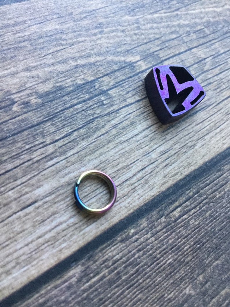 May include: A small, iridescent ring and a purple and black geometric charm rest on a wooden surface. The ring displays a spectrum of colors, while the charm has a cut-out design. The wooden background has a natural, weathered appearance.