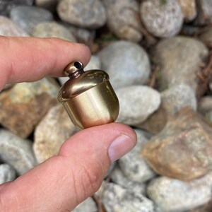 May include: A small, gold-colored acorn pendant. The acorn has a rounded cap and a textured body. The pendant is held between two fingers against a background of smooth, gray and brown stones.