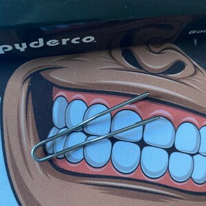 Peut inclure: Une pince à cheveux en métal argenté avec un design simple. La pince est posée sur une surface blanche avec une image de dessin animé d'un visage souriant avec de grandes dents.