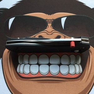 Könnte beinhalten: Ein schwarzes Taschenmesser mit einer silbernen Klinge und einem roten Akzent am Griff. Das Messer ist geschlossen und liegt auf einer weißen Oberfläche mit einem Comic-Affenkopf.