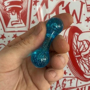 Puede incluir: Un objeto de vidrio azul en forma de pesa con purpurina suspendida en su interior. El objeto tiene una forma redondeada en cada extremo, conectada por un centro cilíndrico más estrecho. El fondo presenta gráficos rojos y blancos.